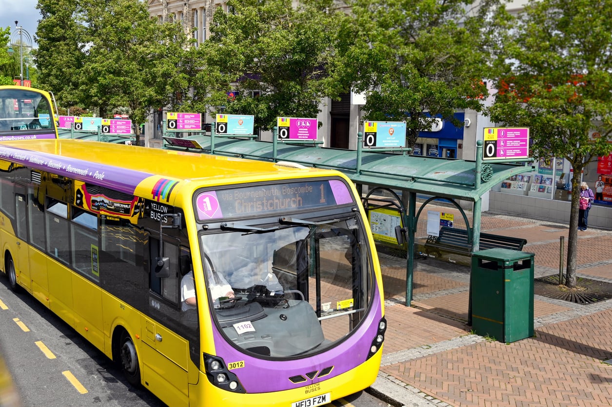 A yellow and purple public bus labeled “Christchurch” stops at a sheltered bus station with green benches and several informational signs, surrounded by trees and brick pavement on a sunny day.