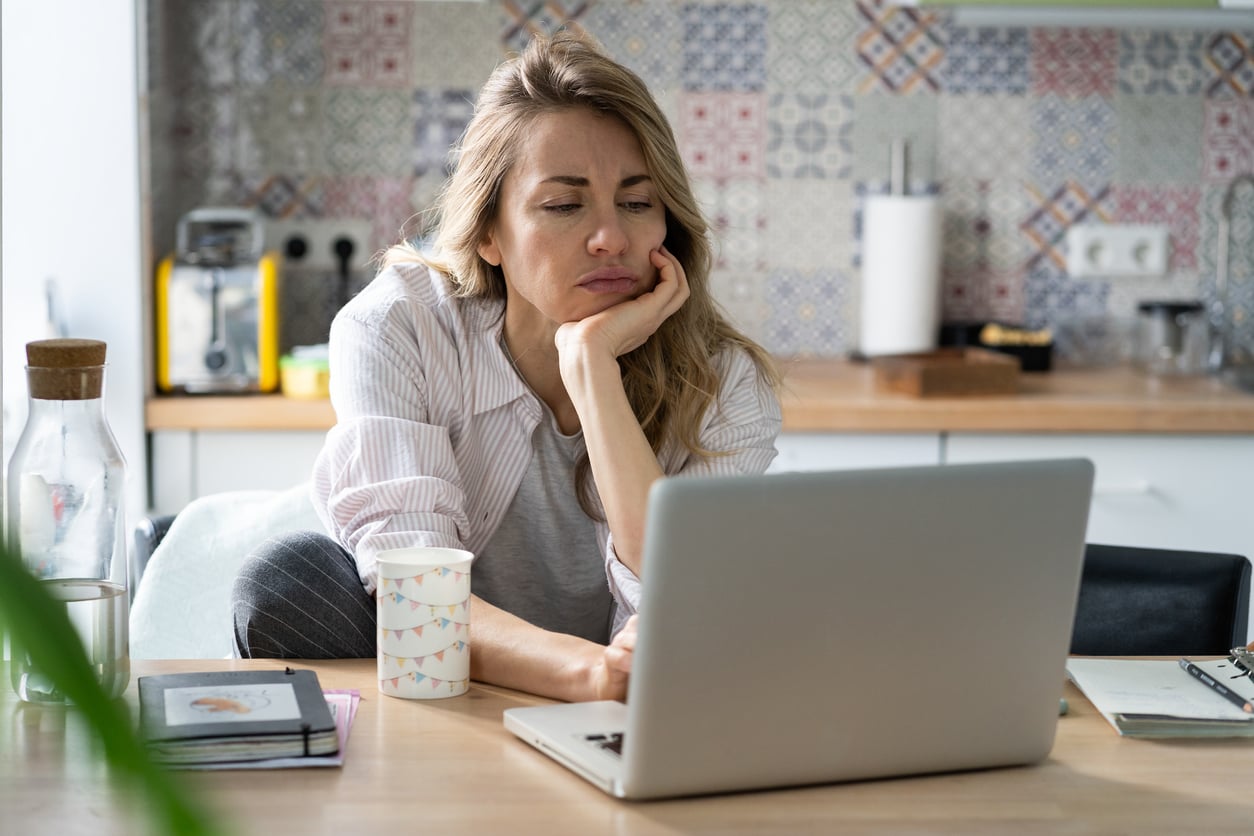 A woman sits at a kitchen table, resting her face on her hand and looking bored or tired while using a laptop—perhaps frustrated by delayed purchases. A cup, water bottle, and notebooks are on the table in front of her.