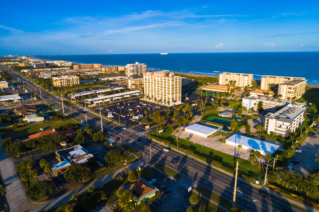 Aerial view of a coastal city with mid-rise buildings, tree-lined streets, and parking lots near a wide sandy beach and blue ocean under a clear sky.