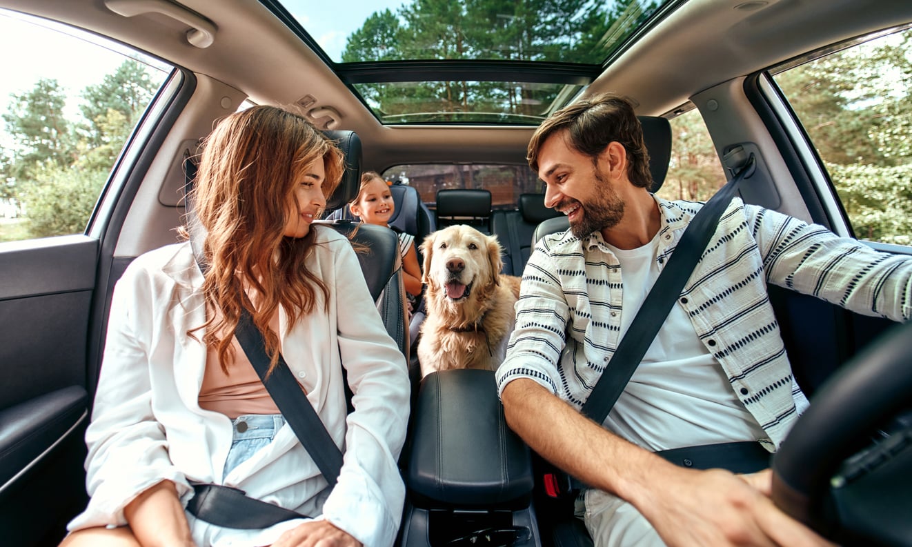 A smiling family, with a woman, a man, a young girl, and a golden retriever dog, sits inside a car with seat belts fastened, driving through a green, tree-filled area on a sunny day.