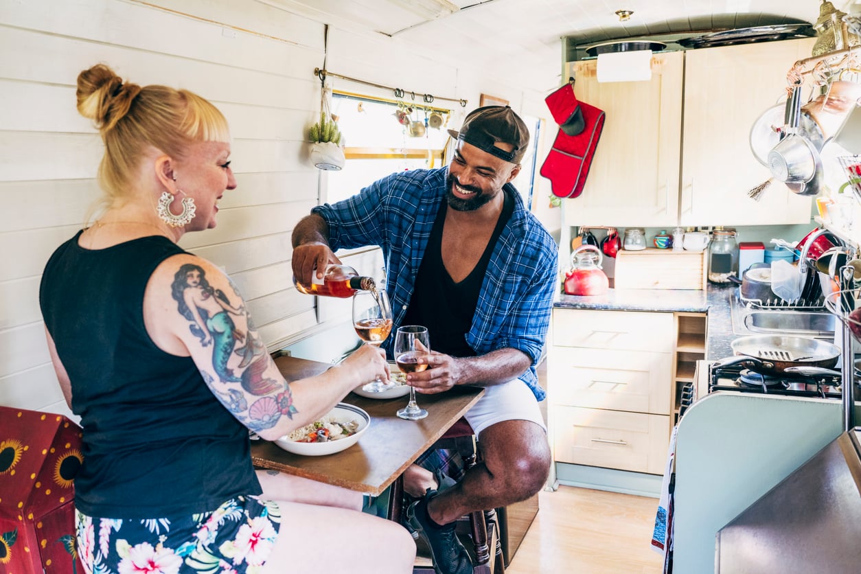 A smiling man pours wine for a smiling woman with tattoos inside a cozy camper van kitchen. They sit at a small table with food, enjoying a casual meal and each other's company.