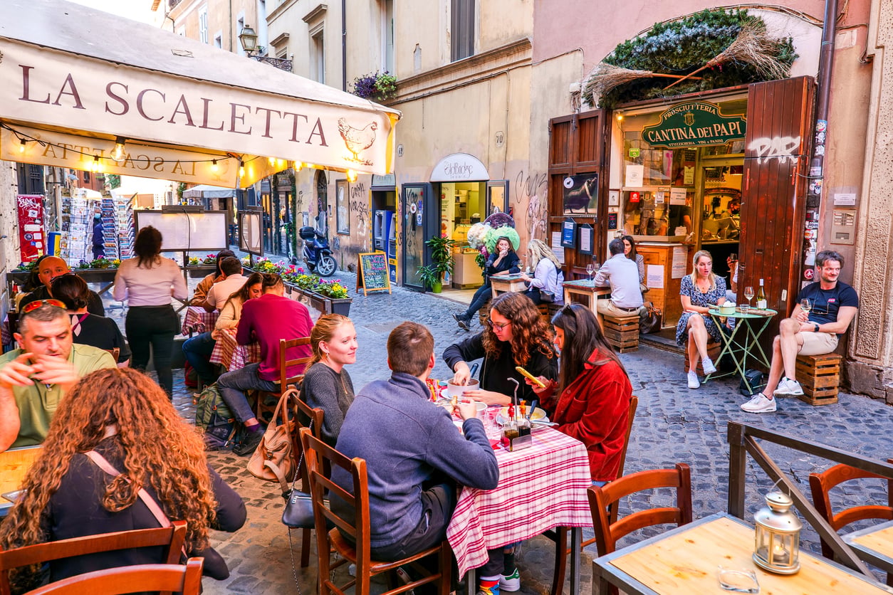 People dine at outdoor tables covered with red and white checkered cloths on a cobblestone street, surrounded by some of the Best Italian Restaurants, creating a lively and colorful European café atmosphere.