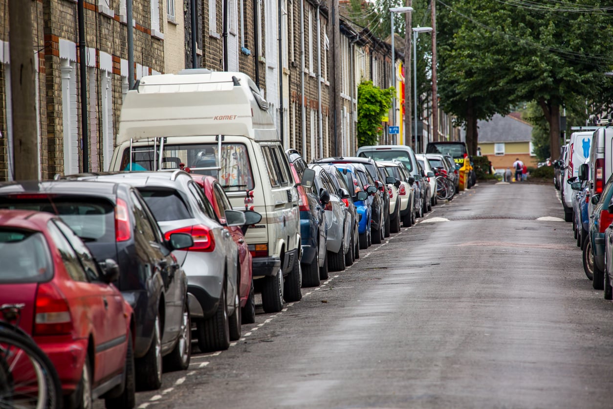 A narrow residential street lined with parked cars on both sides. Brick row houses with white doors are visible on the left, and a few people—perhaps guests staying at an Airbnb—are seen in the distance near the end of the street.