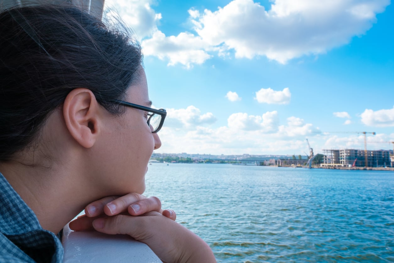 A person wearing glasses rests their chin on their hands while looking out over a calm body of water, reflecting on Florida alternatives beneath a blue sky with clouds and distant buildings.
