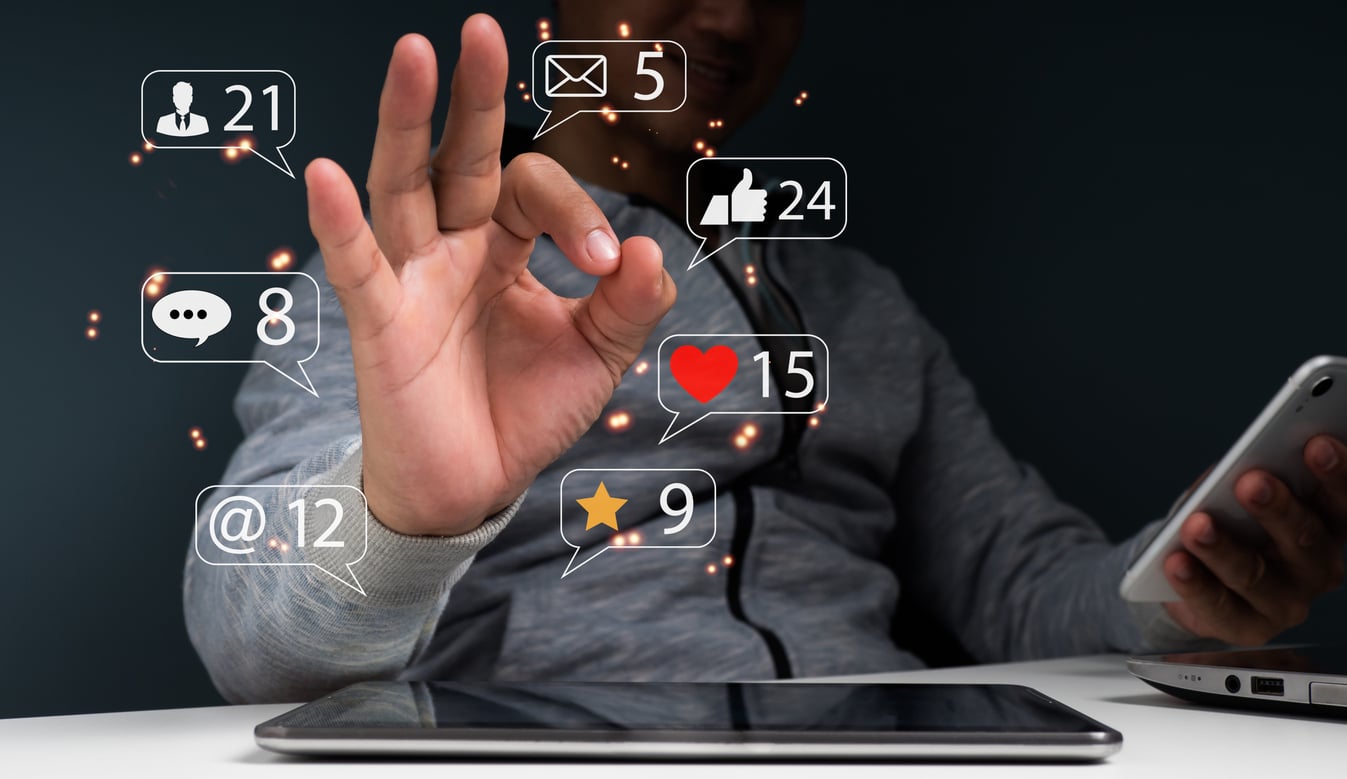 A person in a gray hoodie makes an "OK" gesture near floating social media icons showing notifications for messages, followers, likes, comments, and more, with a tablet and smartphone on the table.
