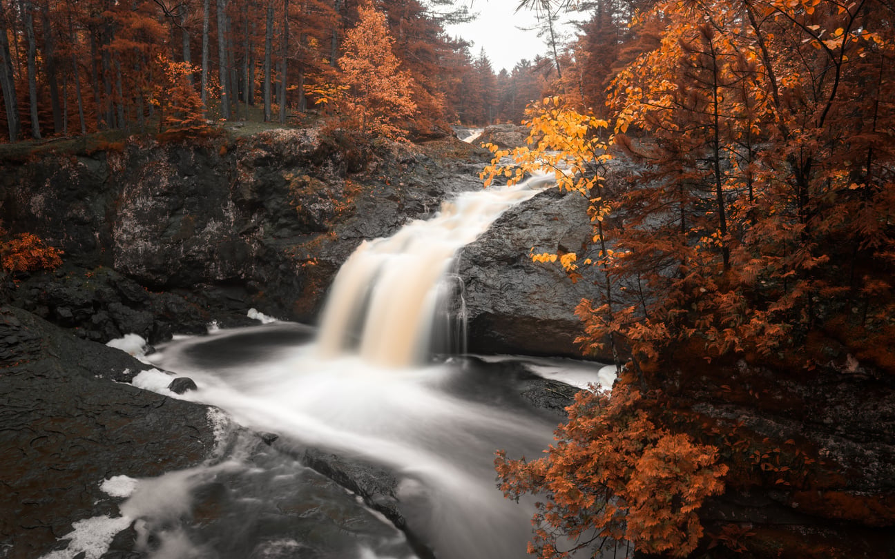 A waterfall cascades over dark rocks surrounded by a forest with vibrant orange autumn leaves, offering a stunning contrast and inviting those seeking Florida alternatives to experience the vivid beauty of fall elsewhere.