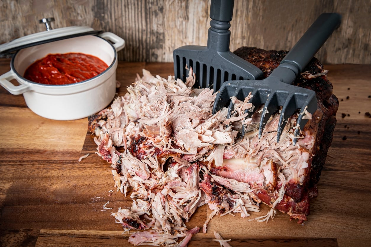Pulled pork being shredded with two meat claws on a wooden surface, next to a white pot of red barbecue sauce. The background features a rustic wooden wall—perfect for those seeking tasty ground beef alternatives.