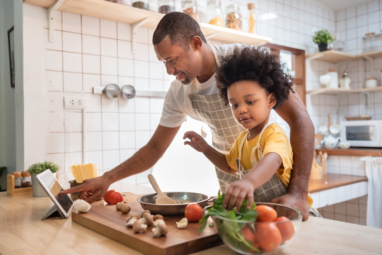 A man and a young child in aprons showcase their DIY skills as they prepare food together in a kitchen. The man touches a tablet, while the child picks vegetables from a glass bowl beside tomatoes and mushrooms on a cutting board.