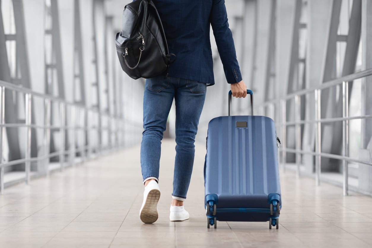 A person in jeans and a blazer walks through an airport corridor, pulling a blue suitcase and carrying a black backpack—ready to put travel hacks to use in the modern setting with sleek glass and metal railings.
