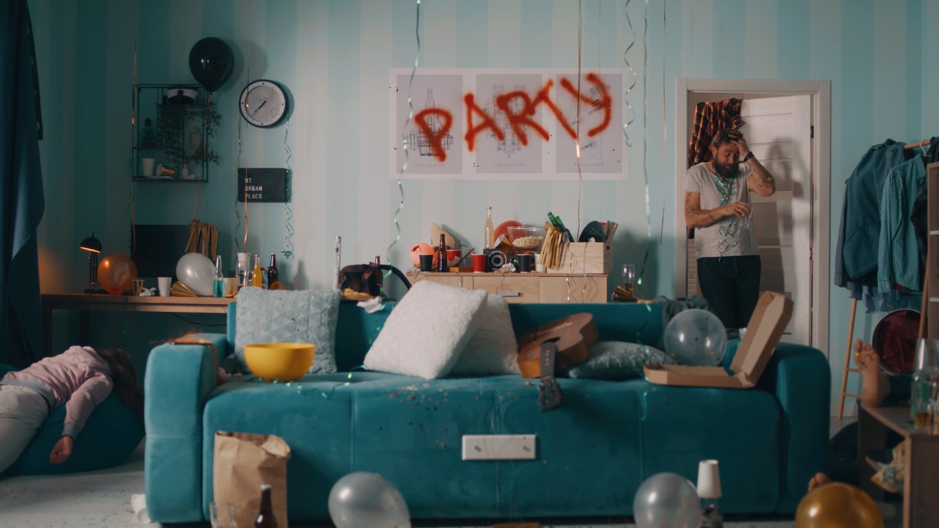 A messy living room after a party at an Airbnb, with balloons, empty bottles, pizza boxes, and spilled snacks. One person lies on the couch while another stands in the doorway. A "PARTY" sign is spray-painted on the wall.