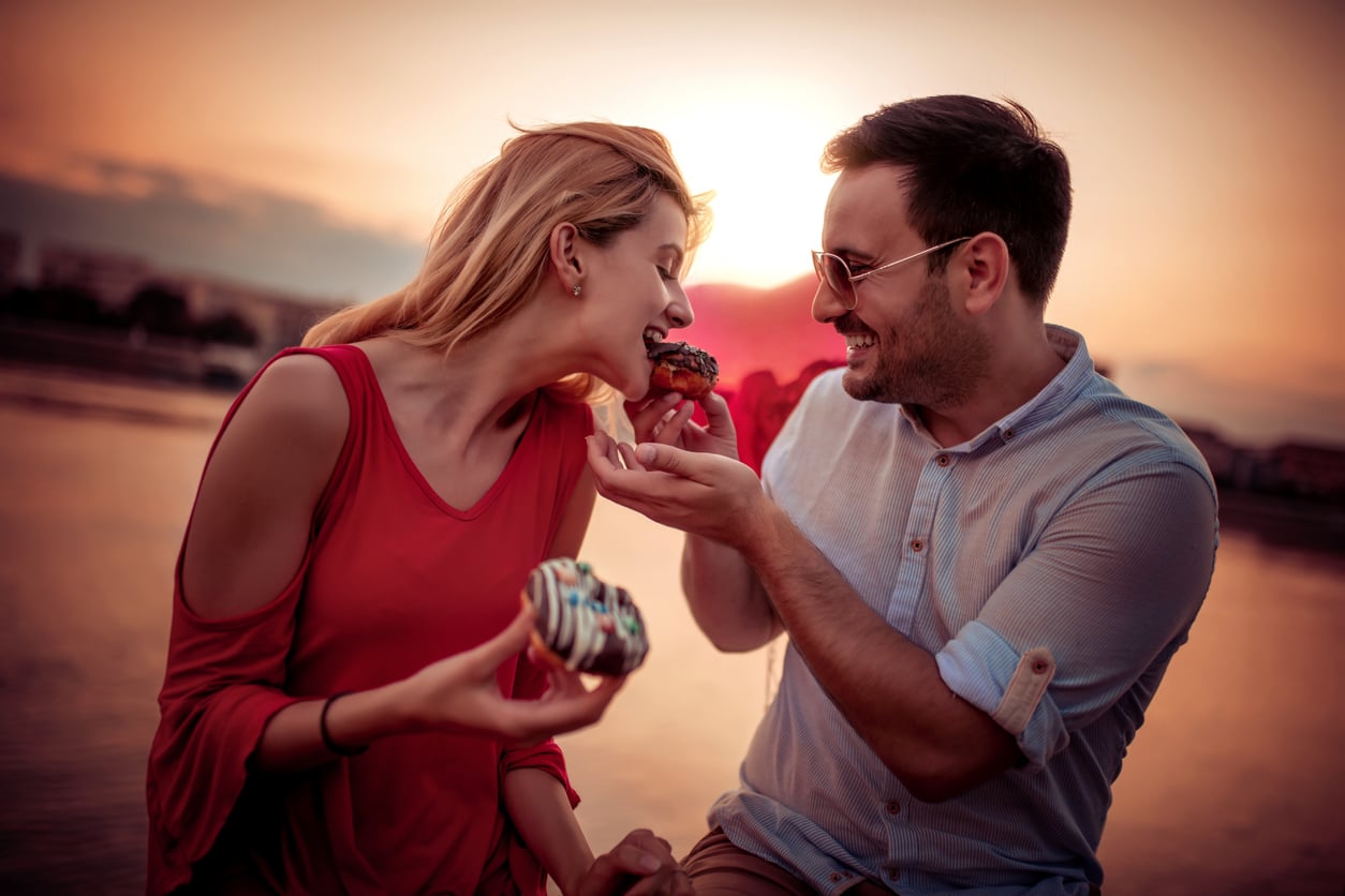 A smiling couple sits by the water at sunset. The man is wearing sunglasses and feeding a donut to the woman, who is holding another donut and leaning in playfully. Both appear happy and relaxed.