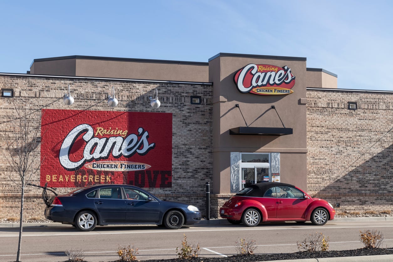 Two cars are parked outside a Raising Cane's Chicken Fingers restaurant with a large mural logo on a brick wall. One car is black and the other is red. The sky is clear and sunny.