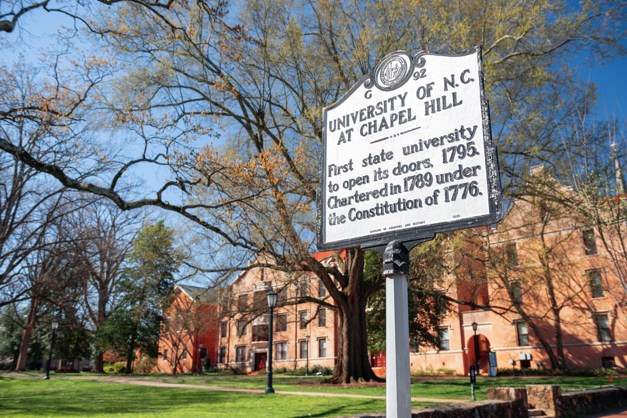 A historical marker for the University of North Carolina at Chapel Hill stands on a grassy lawn, highlighting one of the most affordable college towns, with red-brick campus buildings and tall trees in the background under a clear blue sky.