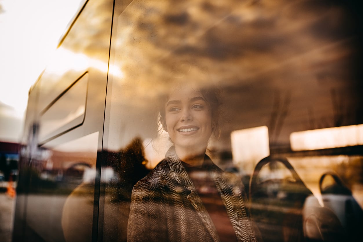 A woman smiles while looking out the window of a bus, with reflections of the cloudy sky and outdoor scenery visible on the glass.