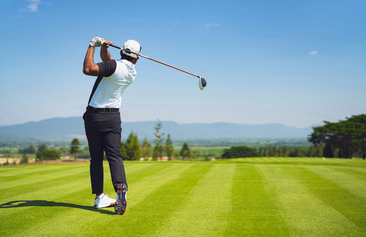 A golfer, embodying the classic style of products men still buy, wears a white shirt and cap as he swings on a well-manicured course, lush green grass and mountains stretching beneath a clear blue sky.
