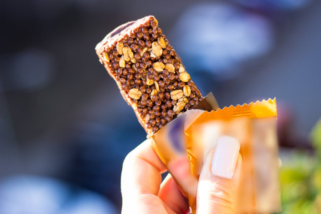 A hand with manicured nails holds a partially unwrapped chocolate-covered granola bar with crispy cereal pieces—perfect for snacking on the go and trying out theme park hacks—against a blurred outdoor background.