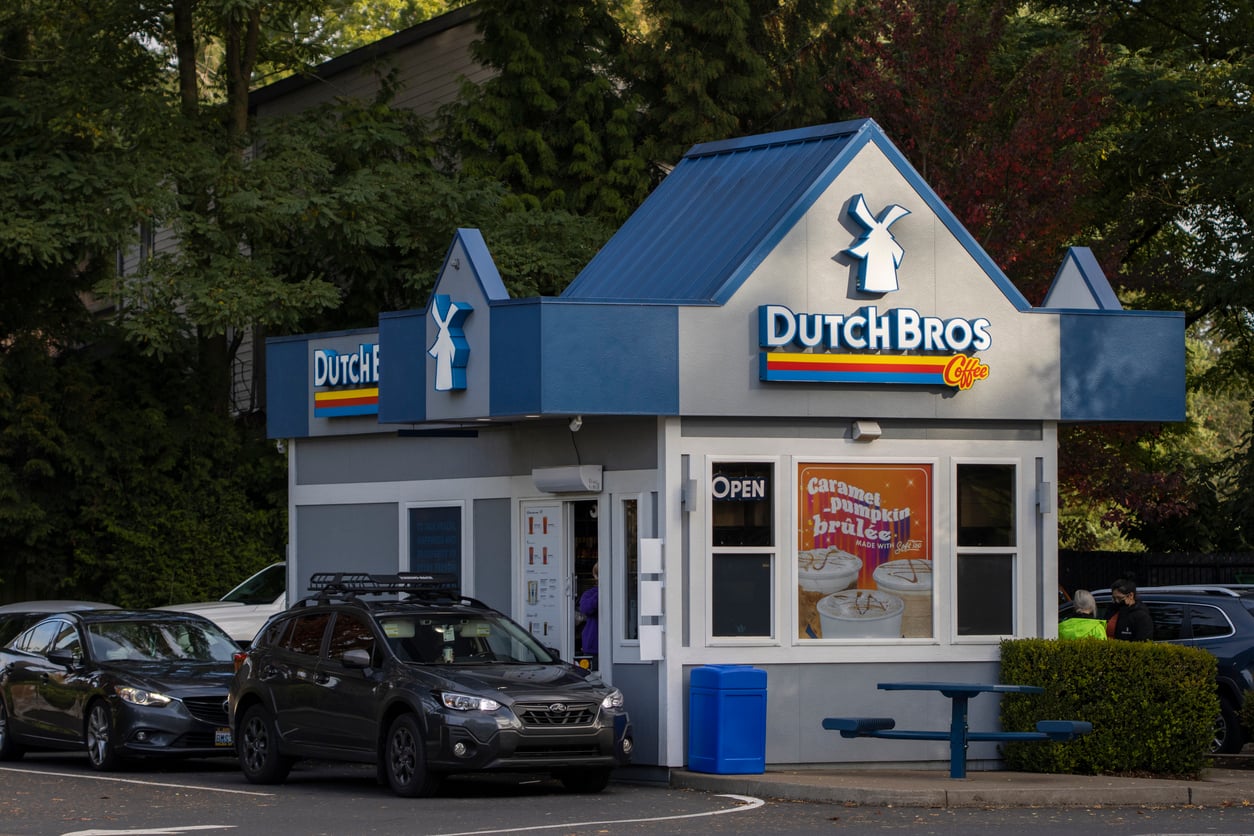 A Dutch Bros Coffee drive-thru shop with blue trim and a windmill logo. Several cars are in line, and colorful signs are visible on the building. Green trees surround the stand.