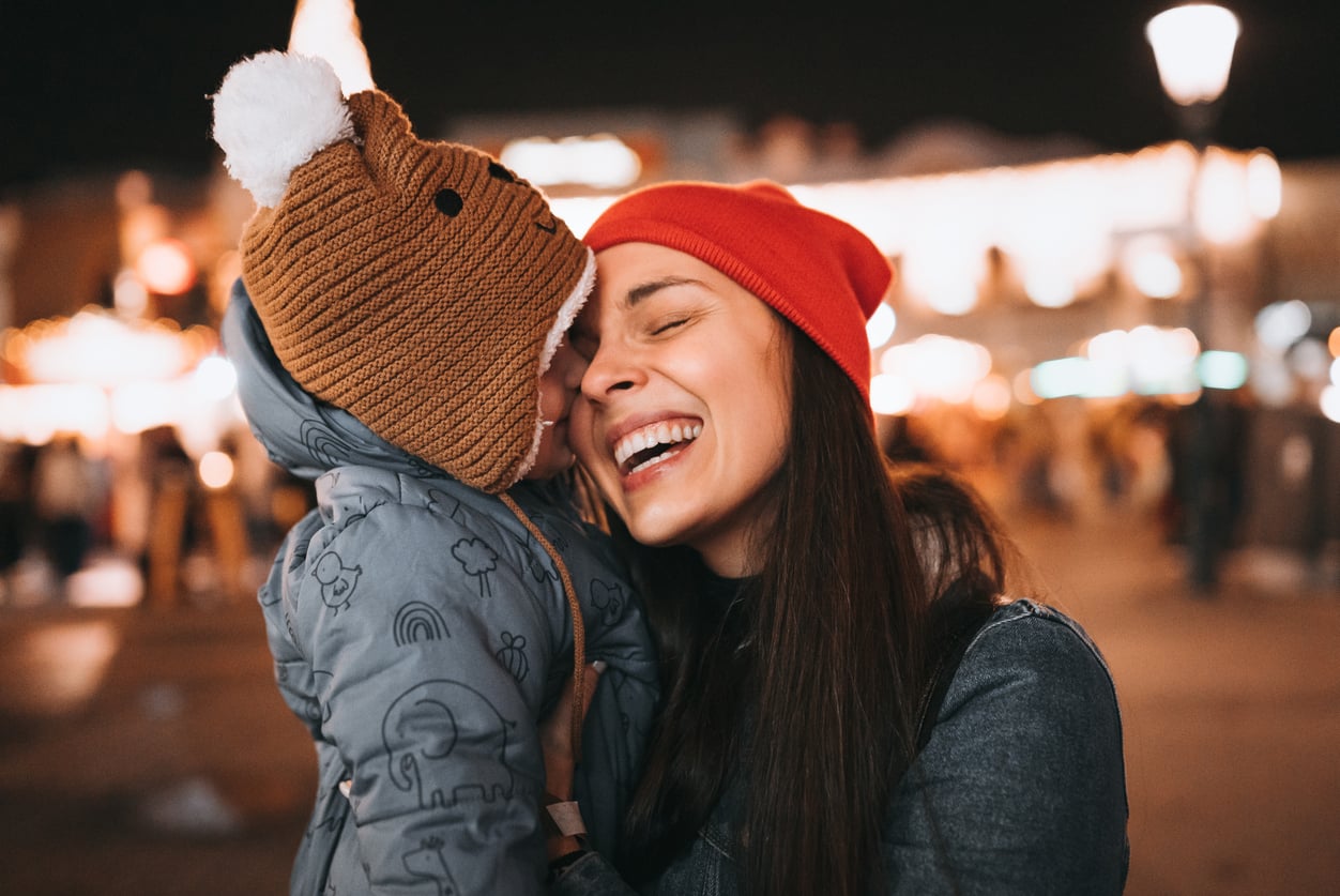 A smiling woman in a red beanie hugs a child in a bear hat and winter jacket at night, surrounded by festive lights and blurred people—capturing the magic of winter evenings and perfect moments for discovering theme park hacks together.