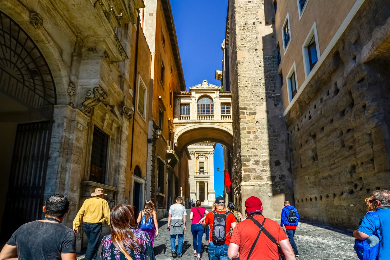 A group of tourists walk down a cobblestone street between tall historic buildings, approaching an ornate arched bridge connecting the structures under a bright blue sky.