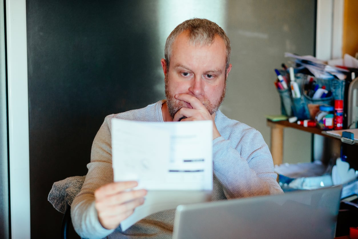 A man with a beard sits at a desk, looking thoughtfully at a document in his hand while working on a laptop. Office supplies and papers are visible on shelves behind him.