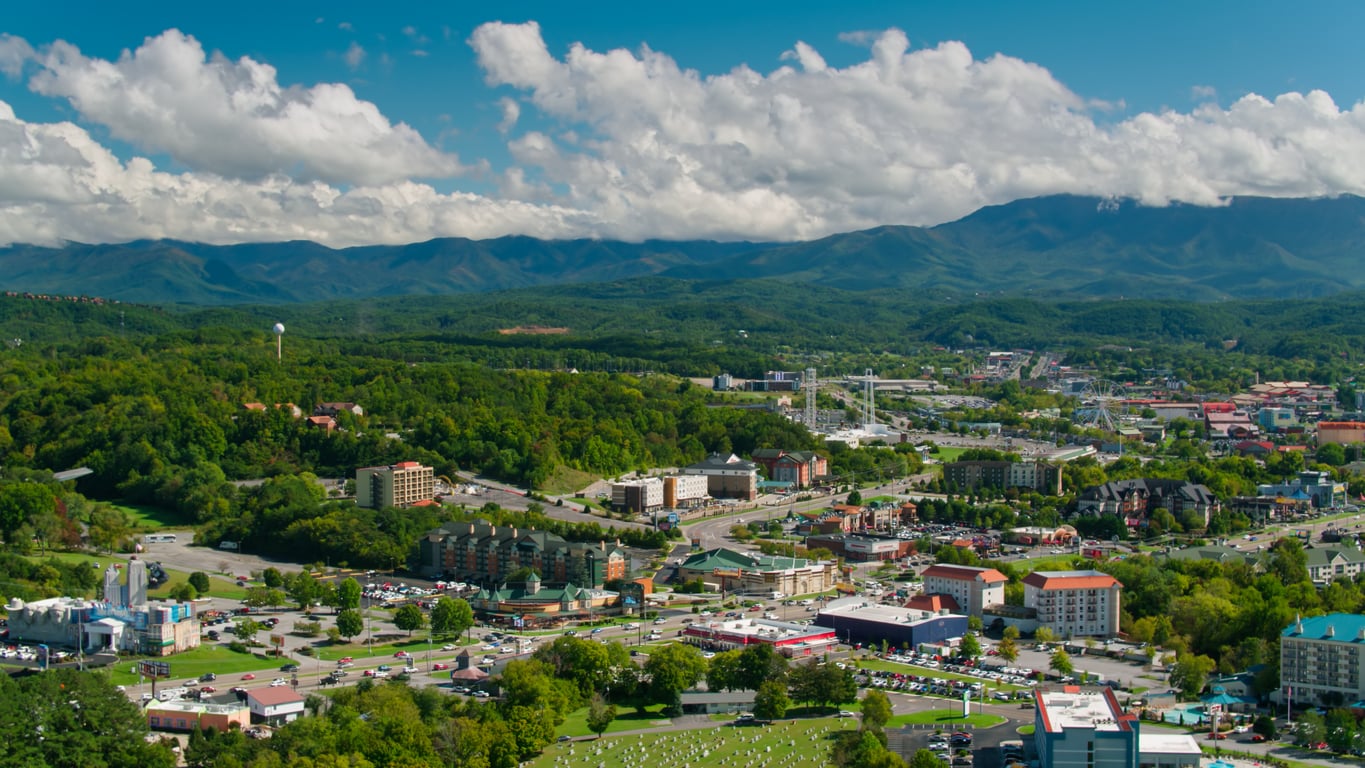 A scenic view of a town surrounded by green trees and rolling hills, with mountains and a partly cloudy sky in the background. Buildings, roads, and cars are visible in the foreground.