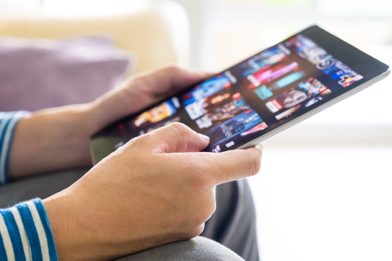 Close-up of a person holding a tablet and browsing through movies or TV shows on a streaming platform, considering which titles to watch without paying extra for overpriced products, with a blurred background and focus on their hands and the device’s screen.