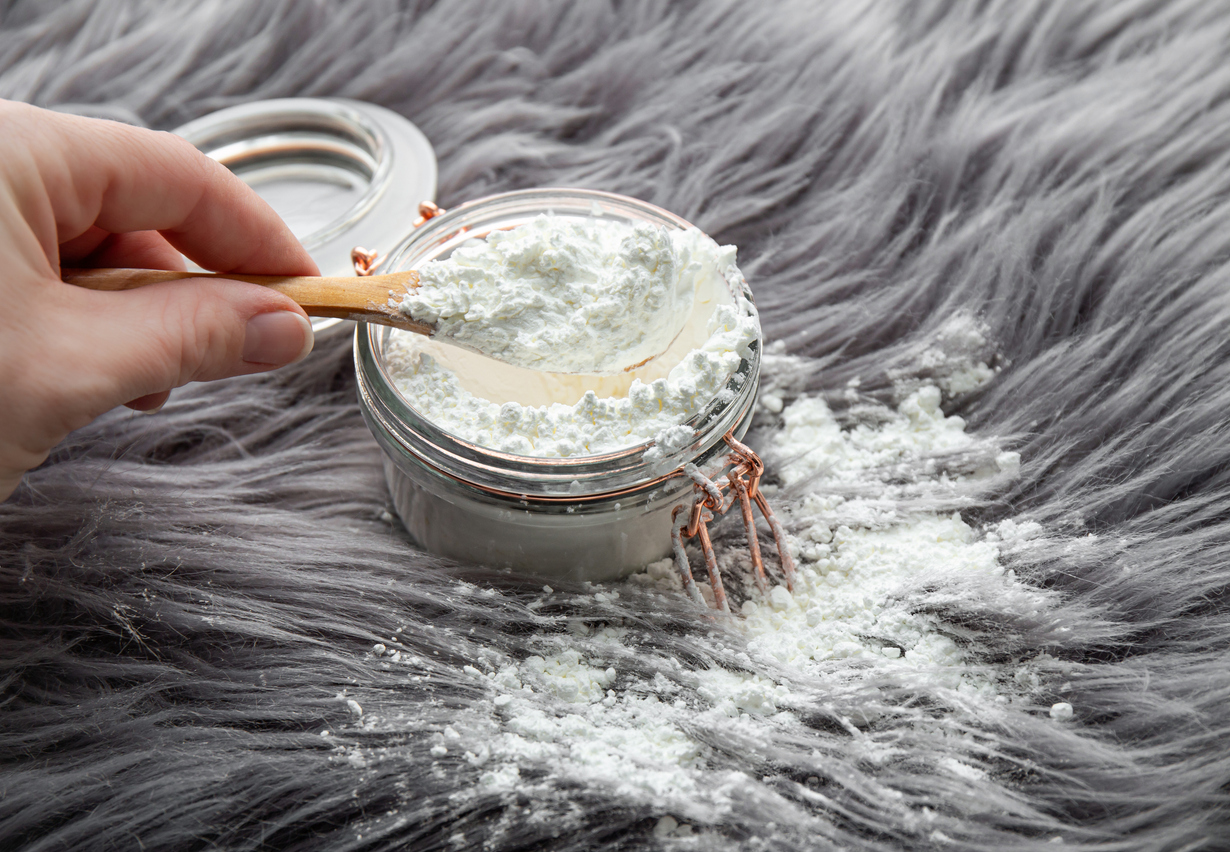 A hand scoops white powder from a glass jar with a wooden spoon—essential for fried chicken tricks. The jar sits on a fluffy gray surface, with some powder spilled beside it.