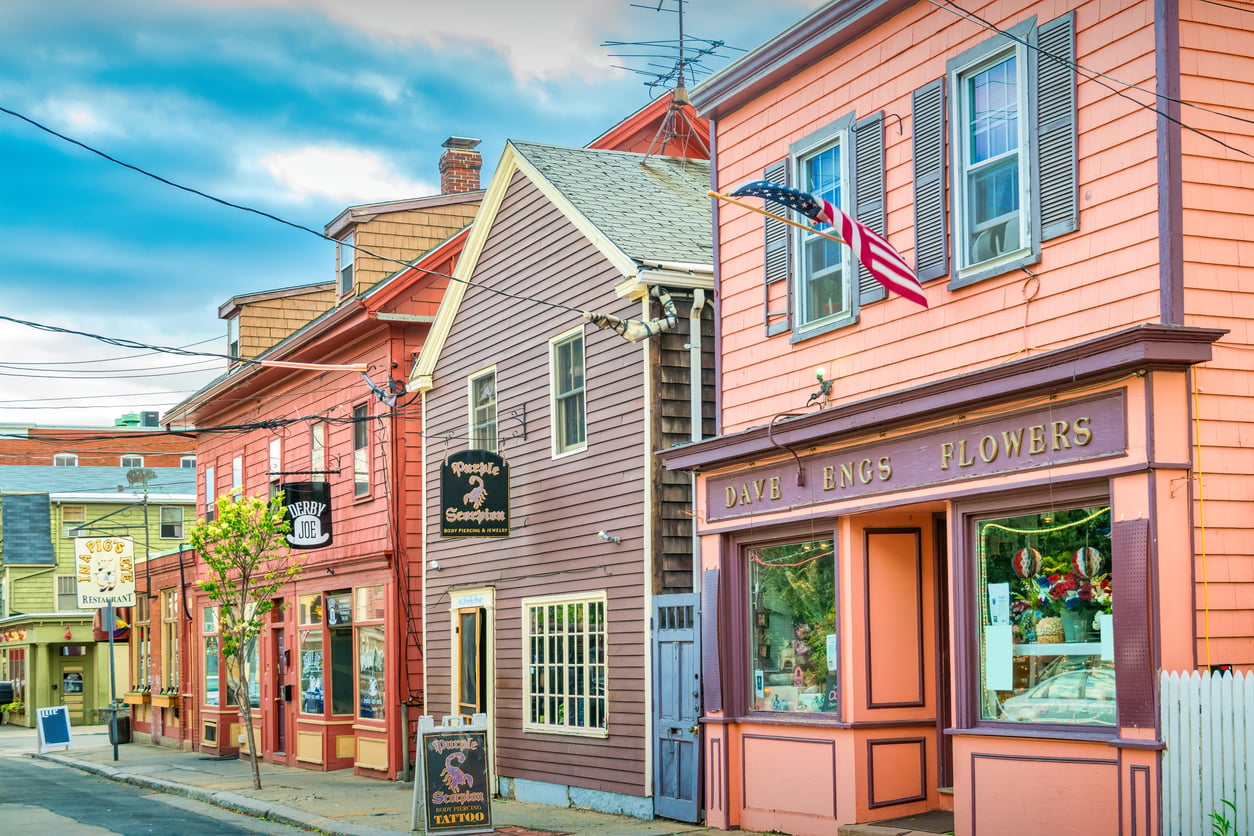 A colorful, small-town street lined with historic wooden buildings showcases the charm of Small Business States, featuring a flower shop and a tattoo parlor beneath an American flag and a bright, partly cloudy sky above.
