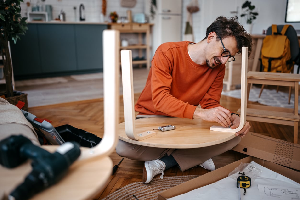 A man in an orange sweatshirt showcases his DIY skills as he sits on the floor, assembling a round wooden table with tools and instructions, surrounded by home furnishings and a toolbox.