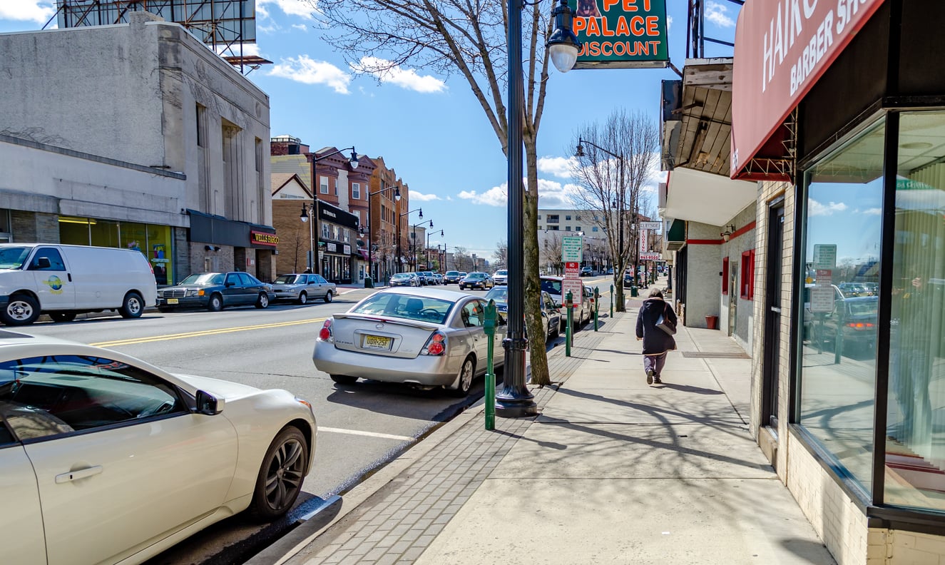 A city street scene with parked cars, shops, and a person walking on the sidewalk. Leafless trees line the street in one of the Small Business States, and the sky is partly cloudy with bright sunlight.