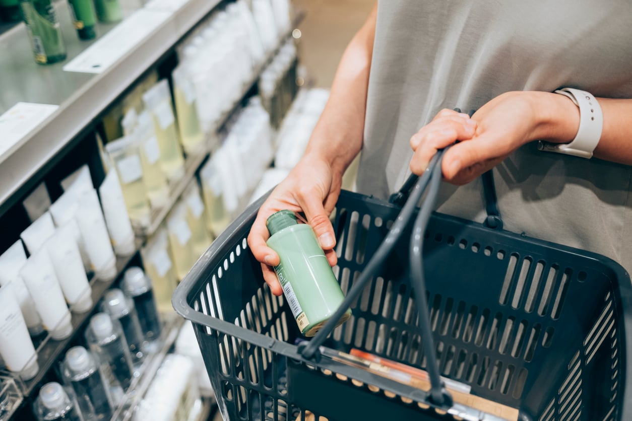 A person holds a green bottle while shopping in a store, carrying a black plastic basket. Shelves stocked with various bottles and containers are visible in the background. Products cheaper online. 