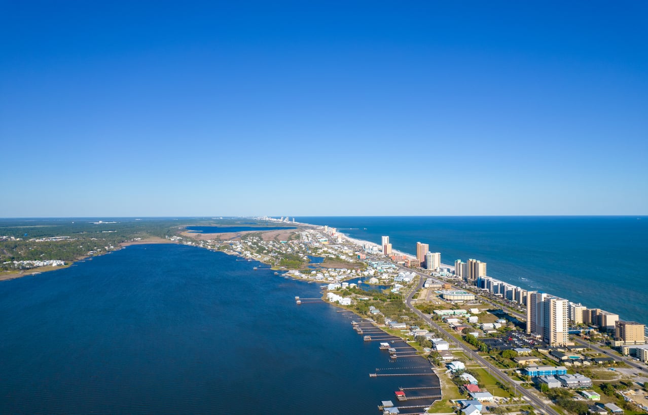 Aerial view of a coastal city with high-rise buildings along a beach, blue ocean on one side, and a large bay on the other, under a clear blue sky—an ideal spot for exploring Florida alternatives.