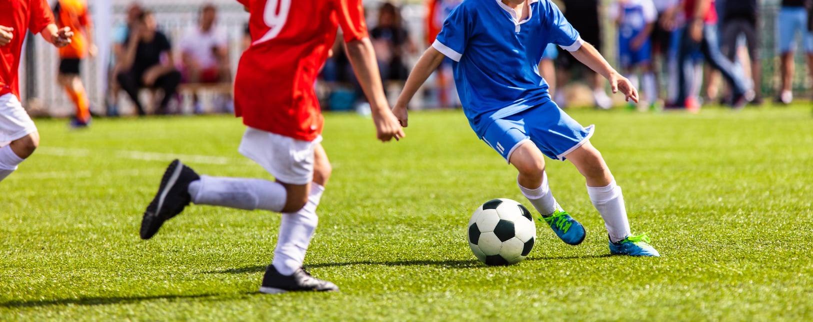 Children playing soccer on a grassy field, with players in blue and red uniforms competing for the ball; only their lower bodies are visible.