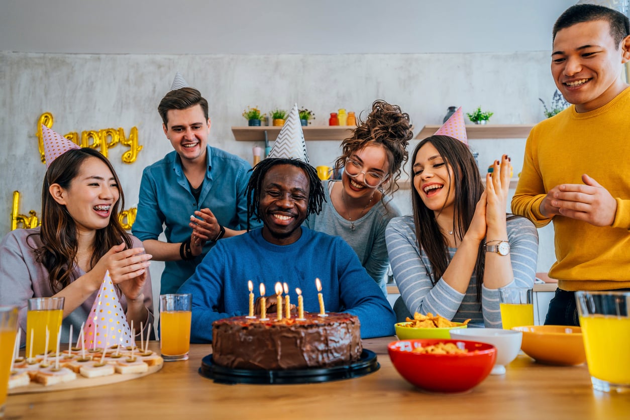 A group of friends in party hats smile and clap around a man seated in front of a chocolate birthday cake with lit candles, celebrating at a table with snacks and drinks.
