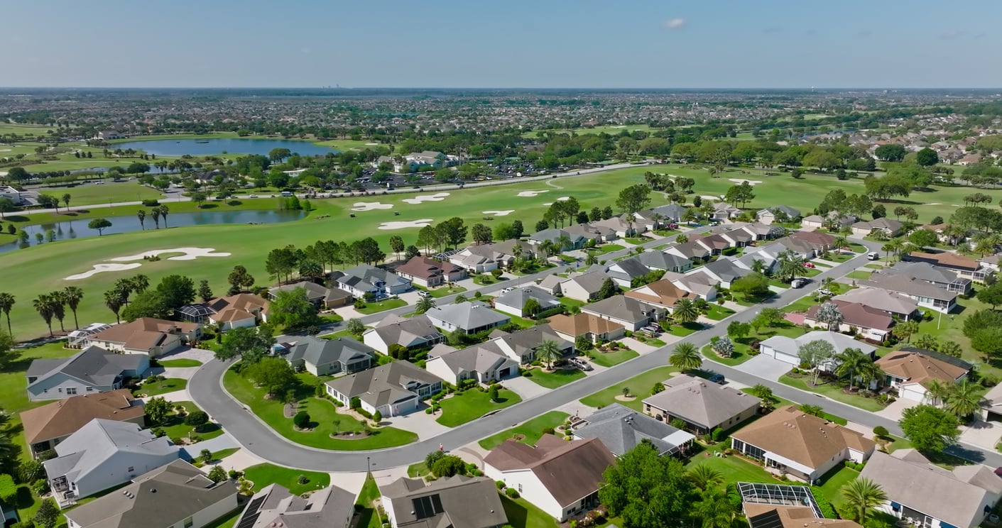 Aerial view of a suburban neighborhood with curving streets, rows of houses with neatly maintained lawns, and a lush green golf course and lakes in the background under a clear blue sky.