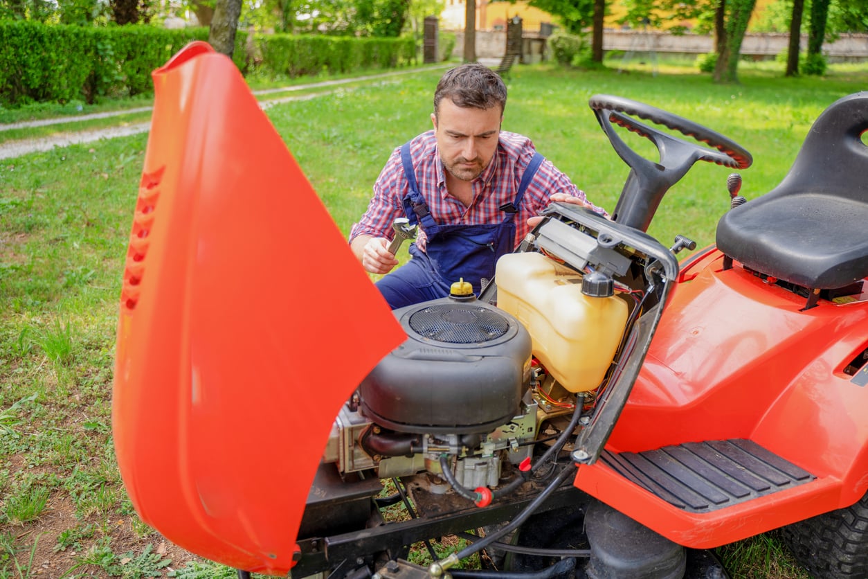 A man in a plaid shirt and overalls demonstrates his DIY skills as he examines the engine of a red riding lawnmower with its hood open, holding a wrench and inspecting the machinery closely in a grassy outdoor area.