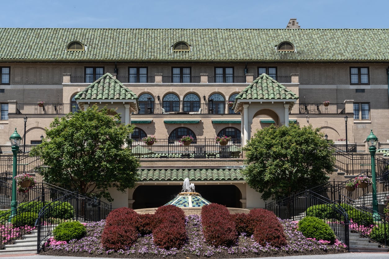 A grand building with a green tiled roof, arched windows, and balconies—perfect for small-town luxury hotels. Trimmed bushes, colorful flowers, two staircases, and a decorative fountain create an inviting entrance.