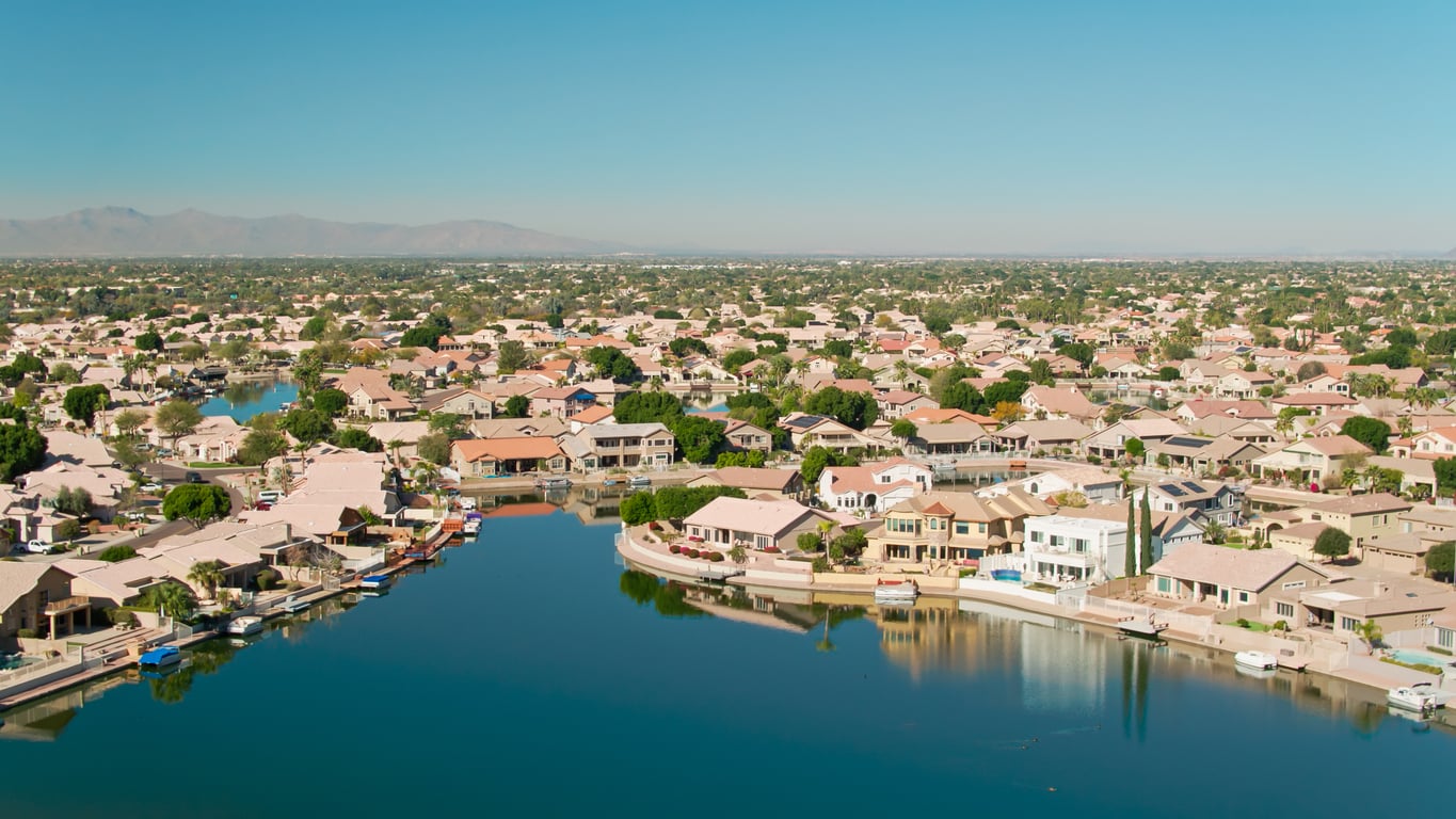 Aerial view of a suburban neighborhood with houses lined along the edge of a calm, blue lake; boats docked at the waterfront, greenery among properties, and mountains visible in the distant background under a clear sky.