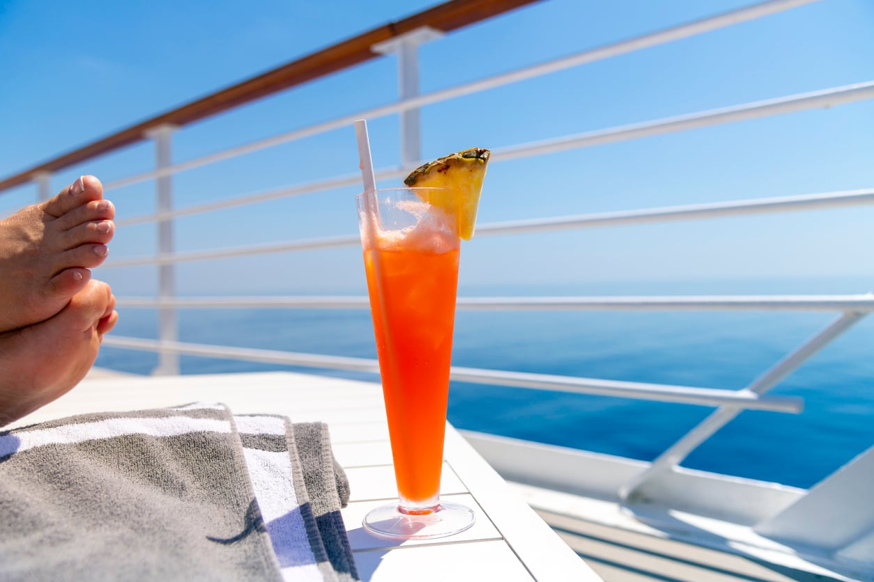 A tropical cocktail with a pineapple slice garnish sits on a table next to a towel, with feet resting nearby and the ocean and clear blue sky visible from a ship's deck—a perfect scene for sharing your favorite travel hacks.