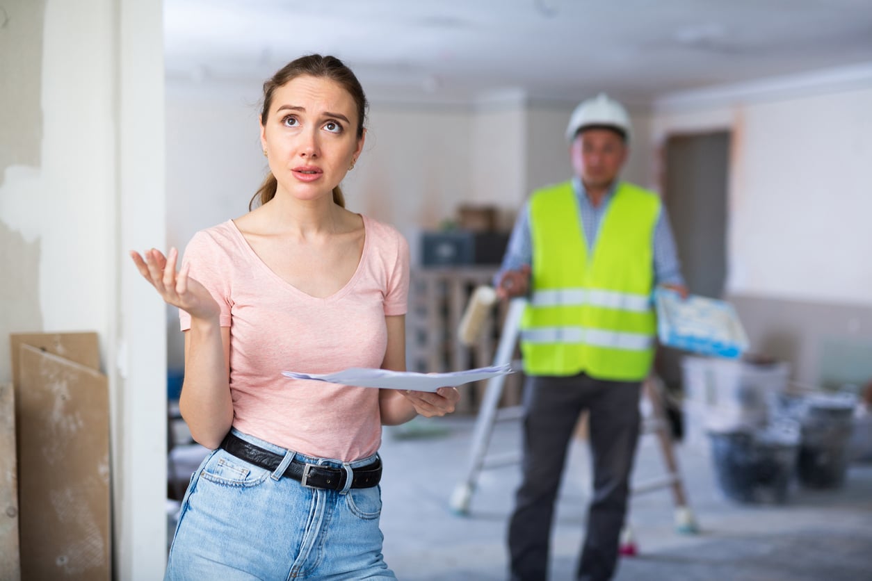 A young woman holding papers looks frustrated—possibly due to delayed purchases—in a room under renovation, while a construction worker in a safety vest and helmet stands in the background with tools.