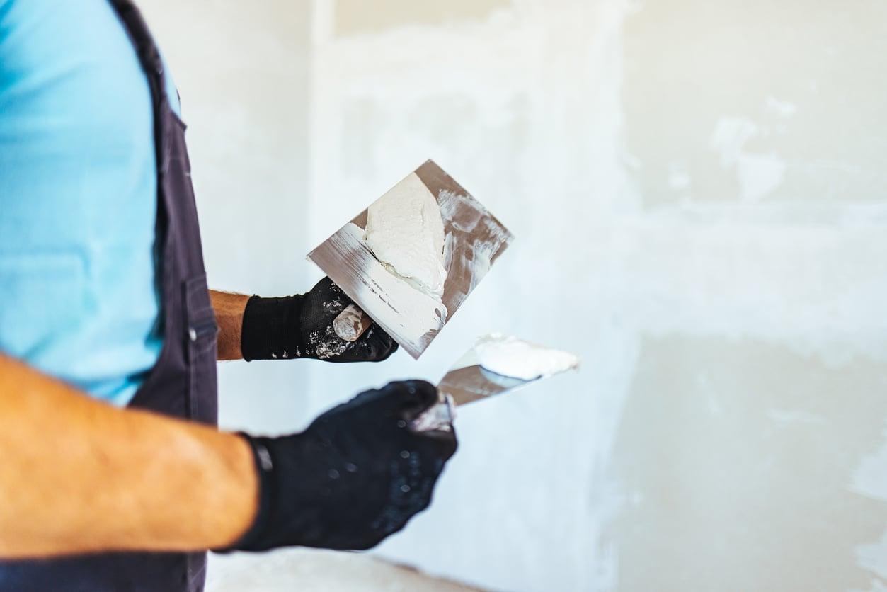A person showcasing their DIY skills wears black gloves and holds two putty knives, one with white plaster, in front of a partially plastered wall.