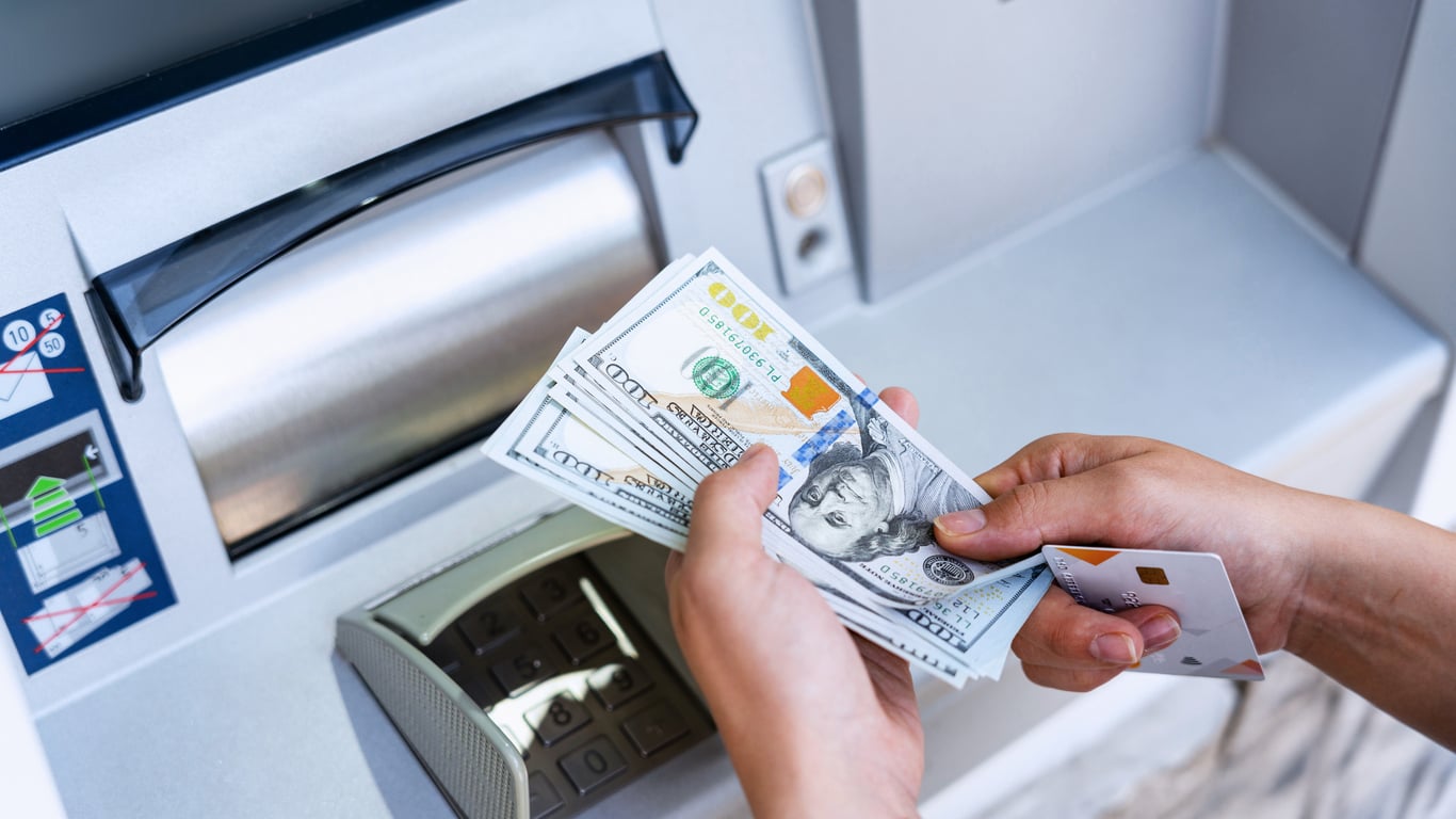 A person holding several US $100 bills and a bank card, standing in front of an ATM machine—ready to put some Vegas money-saving tips into action.