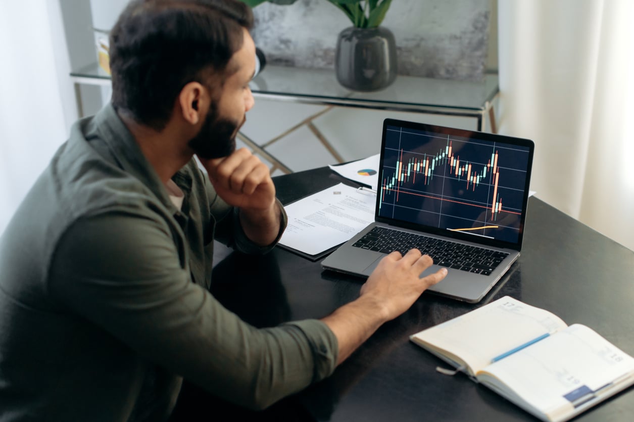 A man with DIY skills sits at a desk, looking at stock market charts on a laptop. Papers and an open notebook are on the table, and a plant is in the background. He appears to be analyzing financial data.
