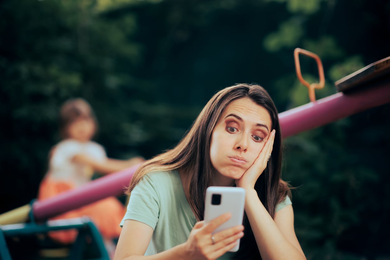 A woman sits outdoors, looking tired and bored as she holds her smartphone in one hand and rests her head on the other. In the blurred background, a child plays on a seesaw. The scene suggests distraction or exhaustion from delayed purchases.
