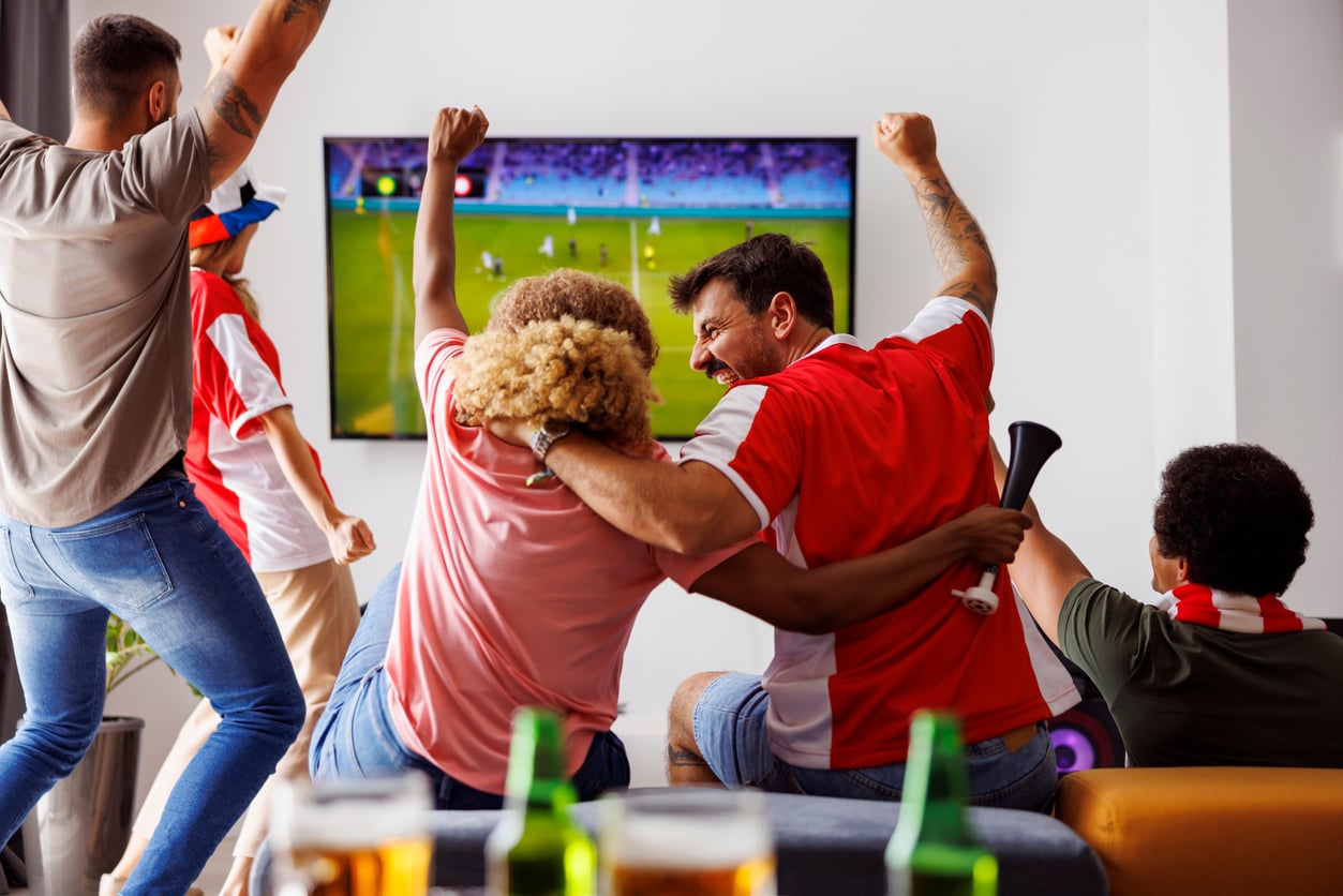 A group of enthusiastic people wearing sports jerseys cheer and celebrate while watching a soccer match on TV in a living room, with classic Products Men Still Buy—like drinks and snacks—on the table in the foreground.