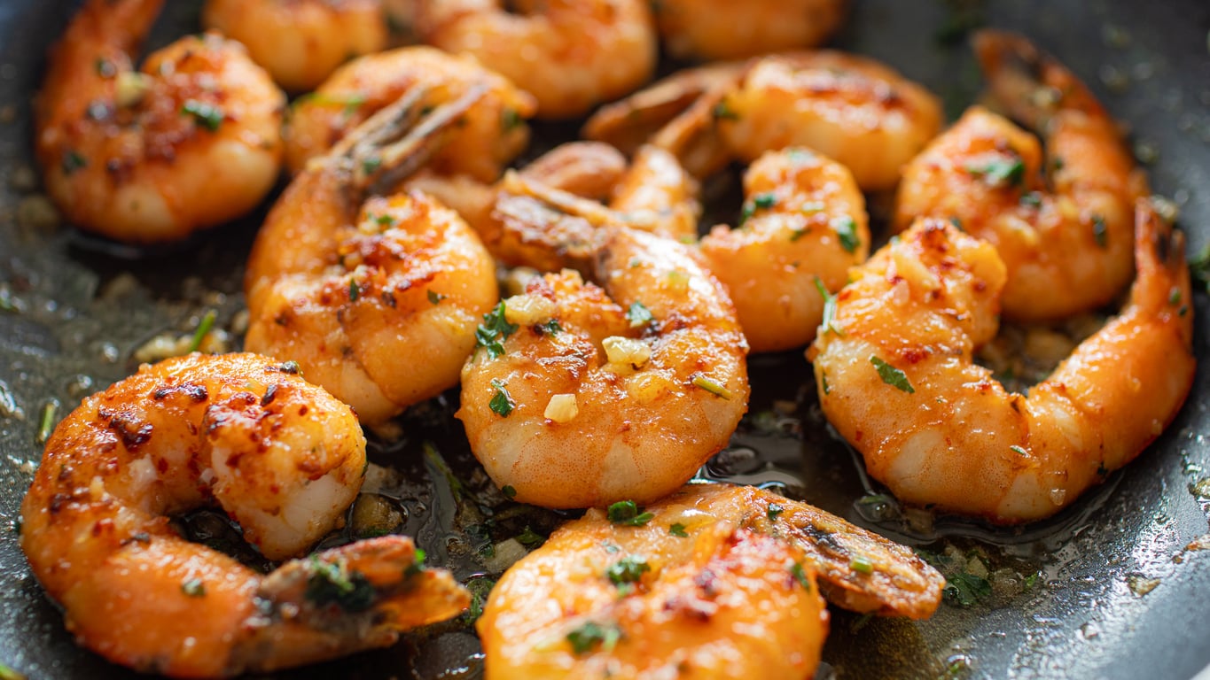 Close-up of cooked shrimp in a pan, seasoned with herbs and spices. The shrimp are orange and appear juicy, with small pieces of garlic and chopped green herbs visible on their surface.