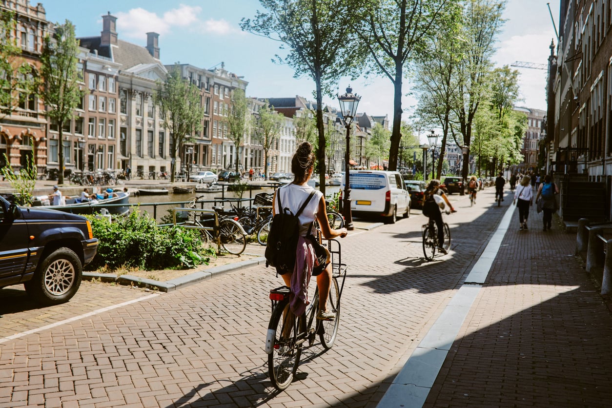 A person rides a bicycle along a sunny canal-side street in a city, with parked cars, more cyclists, pedestrians, and rows of traditional brick buildings in the background. Trees line the street.