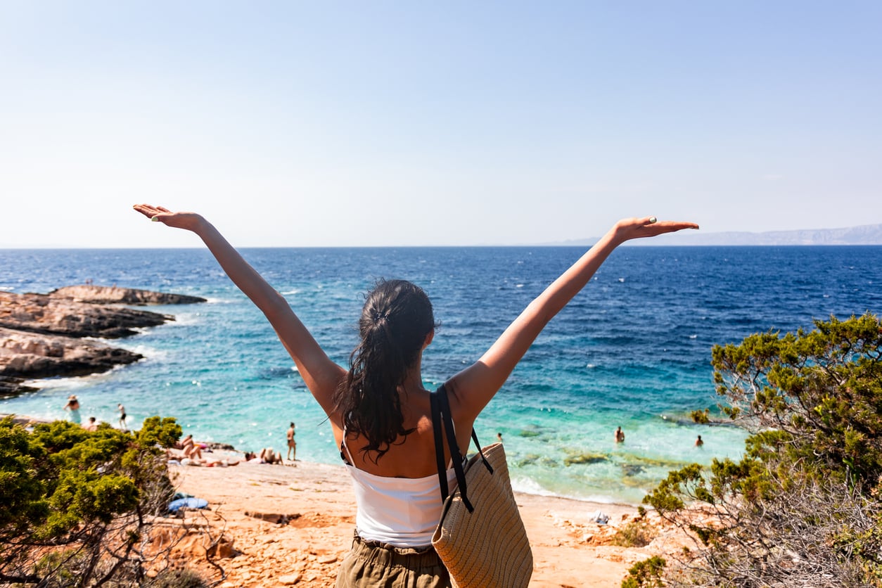 A woman with outstretched arms stands on a rocky shore overlooking a blue sea, with swimmers in the water and greenery nearby, on a sunny day.