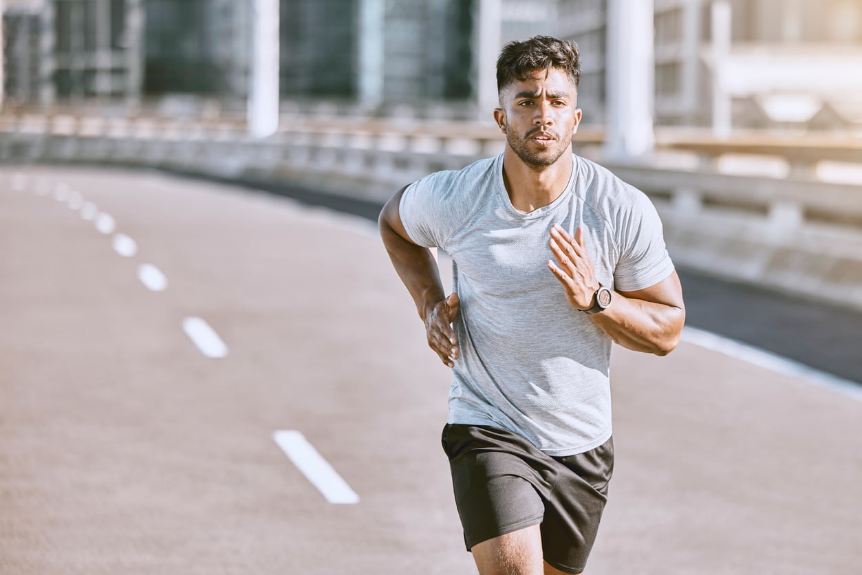 A young man in a light gray t-shirt and black shorts runs on an empty urban road during the day, focused and determined—a perfect example of enjoying budget-friendly hobbies, with city buildings blurred in the background.