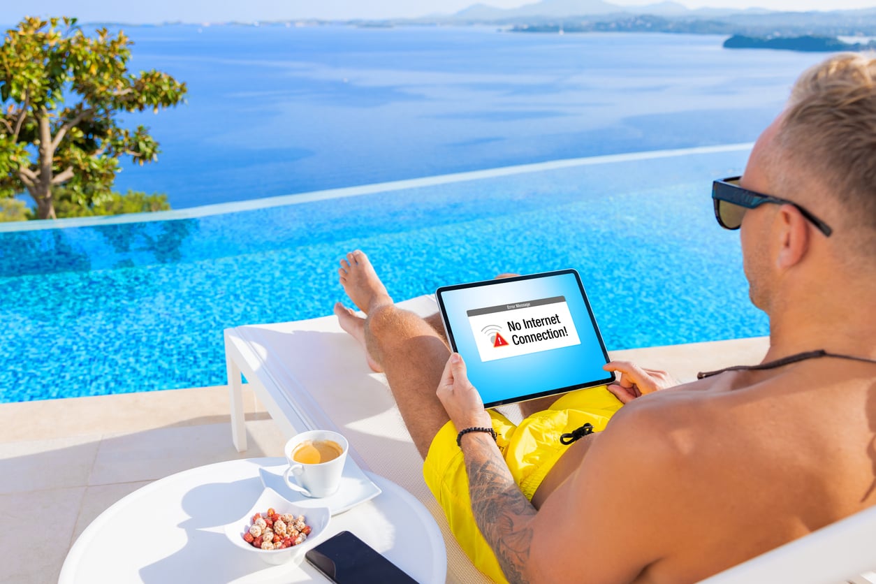 A person in yellow shorts relaxes by a pool with a sea view, staying at an Airbnb, holding a tablet that shows a "No Internet Connection" message. A cup of coffee, snacks, and a smartphone rest on a white table nearby.